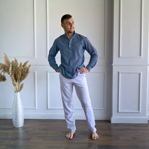 May include: A person wearing a blue striped long-sleeve shirt and white pants. The shirt has a mandarin collar and buttoned cuffs. The pants are a light color and have a relaxed fit. The person is standing on a wooden floor.