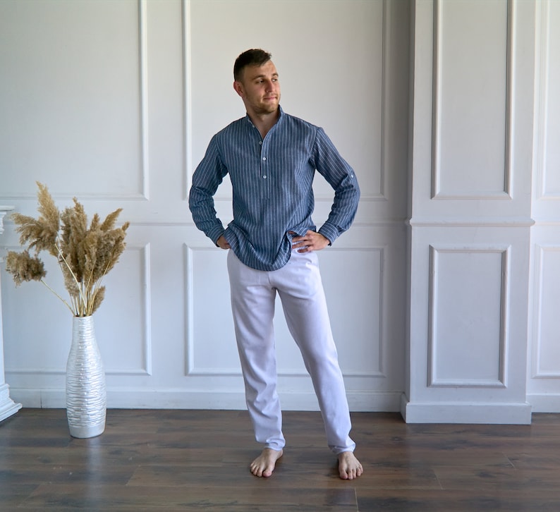 May include: A person wearing a blue striped long-sleeve shirt and white pants. The shirt has a mandarin collar and buttoned cuffs. The pants are a light color and have a relaxed fit. The person is standing on a wooden floor.