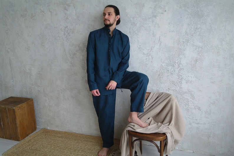 Pode incluir: Uma pessoa vestindo uma camisa de manga comprida azul escura e cal&ccedil;as combinando. A camisa tem uma gola mandarim e fechamento com bot&otilde;es. A pessoa est&aacute; em p&eacute; com uma perna levantada e apoiada em uma cadeira de madeira com um cobertor bege drapeado sobre ela.