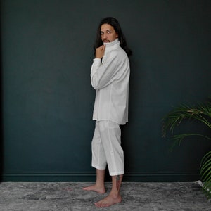 May include: A white collared shirt with long sleeves and a white pair of pants. The person is standing in front of a dark green wall.