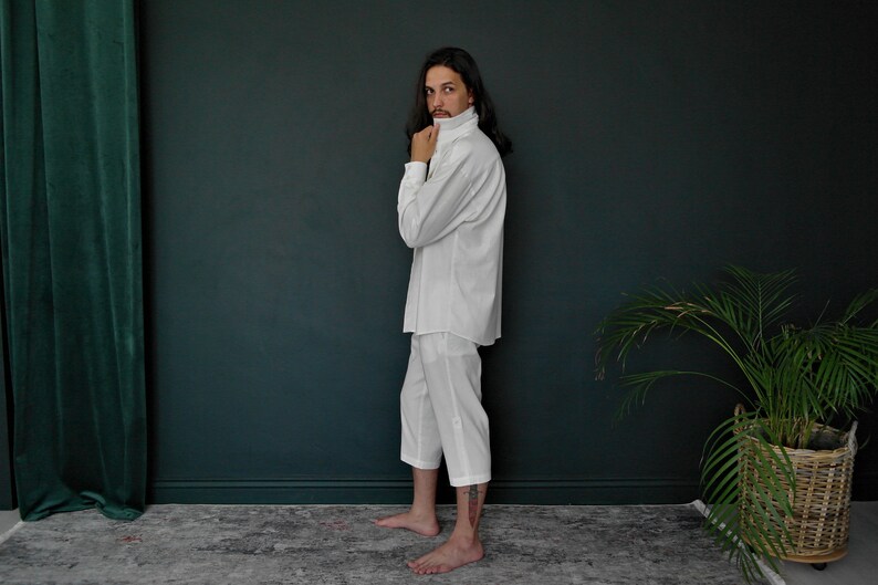 May include: A white collared shirt with long sleeves and a white pair of pants. The person is standing in front of a dark green wall.