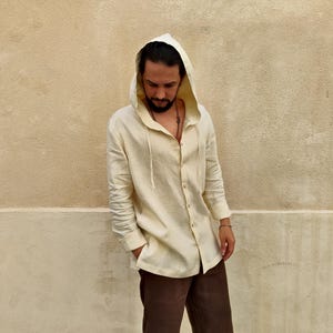 Può includere: Una camicia di lino color crema con cappuccio e maniche lunghe. La camicia ha una chiusura con bottoni sul davanti e un cappuccio con coulisse.
