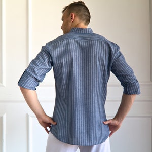 May include: A blue and white striped shirt with long sleeves, rolled up at the elbows. The shirt has a mandarin collar and is buttoned down the front. The model is wearing white pants. The shirt is made of a lightweight fabric.