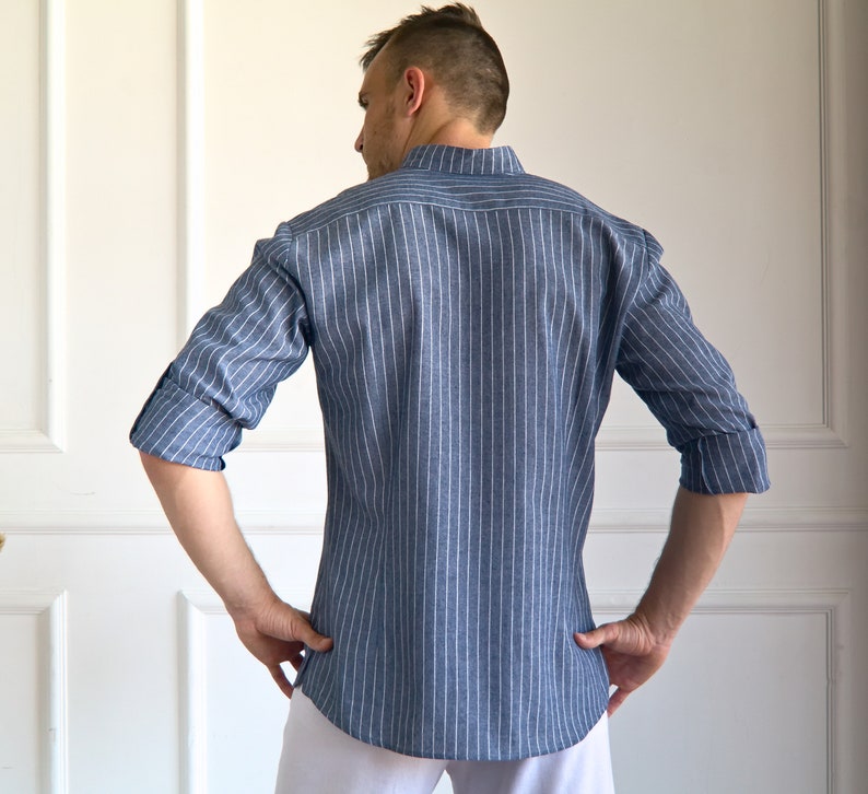 May include: A blue and white striped shirt with long sleeves, rolled up at the elbows. The shirt has a mandarin collar and is buttoned down the front. The model is wearing white pants. The shirt is made of a lightweight fabric.