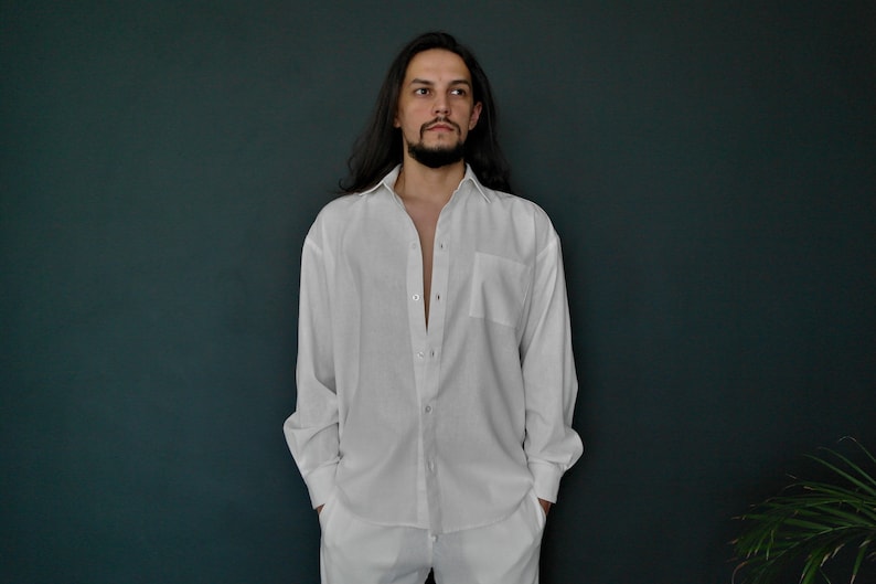 May include: A man with long dark hair is wearing a white button-down shirt with long sleeves. He is standing in front of a dark green wall.