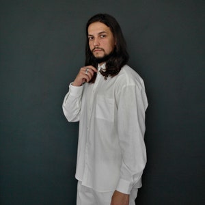 May include: A man wearing a white long-sleeved button-down shirt with a collar. He is standing in front of a dark green wall.