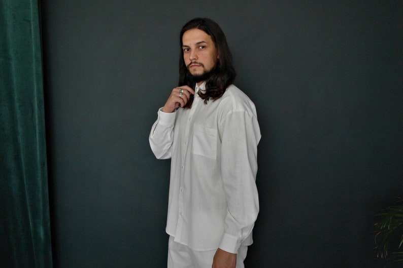 May include: A man wearing a white long-sleeved button-down shirt with a collar. He is standing in front of a dark green wall.