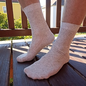 Pode incluir: Um par de meias bege claro com uma textura mesclada subtil e um padrão de malha decorativo vertical ao longo do lado de cada meia. As meias têm um punho canelado na parte superior e uma zona reforçada na biqueira e no calcanhar.