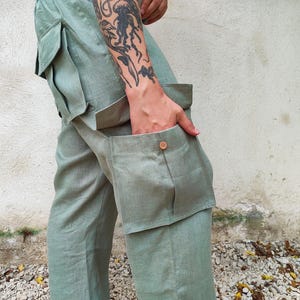 Puede incluir: Pantalones cargo verde claro con bolsillos laterales grandes y un detalle de botón. Los pantalones están hechos de una tela ligera y tienen un ajuste relajado. La persona lleva los pantalones con la mano en el bolsillo.