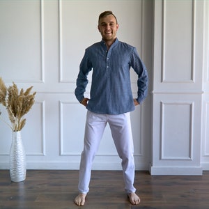 May include: A person wearing a blue striped long-sleeve shirt and white pants. The shirt has a mandarin collar and a buttoned placket. The pants are a solid color and have a straight leg. The person is standing in front of a white wall.