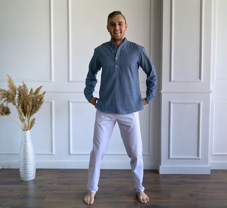 May include: A person wearing a blue striped long-sleeve shirt and white pants. The shirt has a mandarin collar and a buttoned placket. The pants are a solid color and have a straight leg. The person is standing in front of a white wall.
