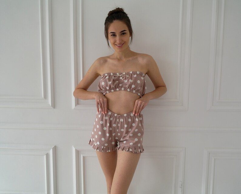 May include: A woman is wearing a pink and white polka dot pajama set. The set includes a strapless top and shorts with a ruffled hem.