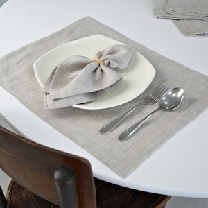 Puede incluir: Un mantel individual de lino gris con un plato cuadrado blanco, una servilleta de lino doblada en forma de lazo y cubiertos de plata.