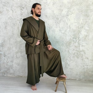 May include: A long, olive green robe with a hood and a sash. The robe is worn over a dark green shirt. The person is standing with one leg raised on a wooden stool.