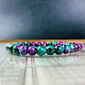 Puede incluir: Un brazalete de cuentas redondas pulidas, en tonos morados, turquesas y verdes. Las cuentas están ensartadas para formar un círculo. El brazalete se muestra sobre una superficie reflectante, con un fondo oscuro.