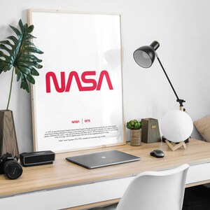 May include: A framed poster with a white background and the red NASA logo in a bold, sans-serif font. The text "NASA 1975" is printed below the logo in a smaller font. The poster is hanging on a white wall above a wooden desk with a laptop, a lamp, and a potted plant.