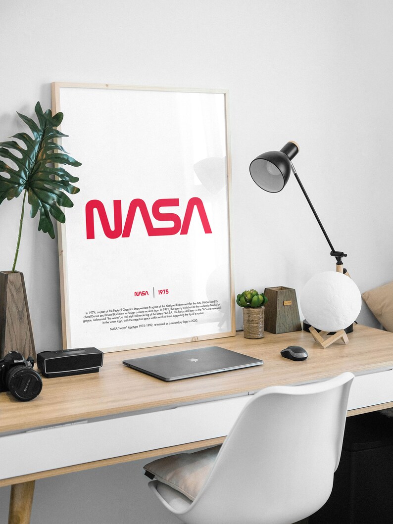 May include: A framed poster with a white background and the red NASA logo in a bold, sans-serif font. The text "NASA 1975" is printed below the logo in a smaller font. The poster is hanging on a white wall above a wooden desk with a laptop, a lamp, and a potted plant.