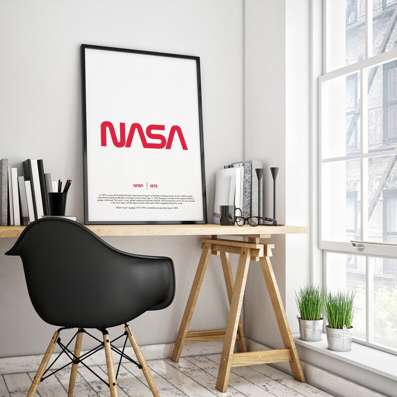 May include: A black framed poster with a red NASA logo and the text "NASA | 1975" on a white background. The poster is hanging on a white wall above a desk with a black chair, a wooden easel, and a potted plant.