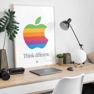 May include: A framed poster with a rainbow apple logo and the text "Think different." The poster is hanging on a white wall above a wooden desk with a laptop, a lamp, and a white chair.