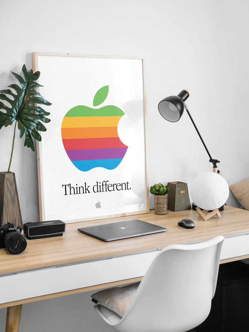 May include: A framed poster with a rainbow apple logo and the text "Think different." The poster is hanging on a white wall above a wooden desk with a laptop, a lamp, and a white chair.