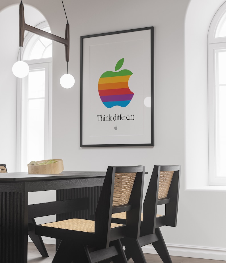 May include: A black framed poster with a rainbow colored Apple logo and the text "Think different." The poster is hanging on a white wall above a black dining table with two black chairs with woven cane seats.