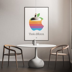 May include: A white poster with a rainbow apple logo and the text "Think different." hanging on a white wall above a white table with two wooden chairs.