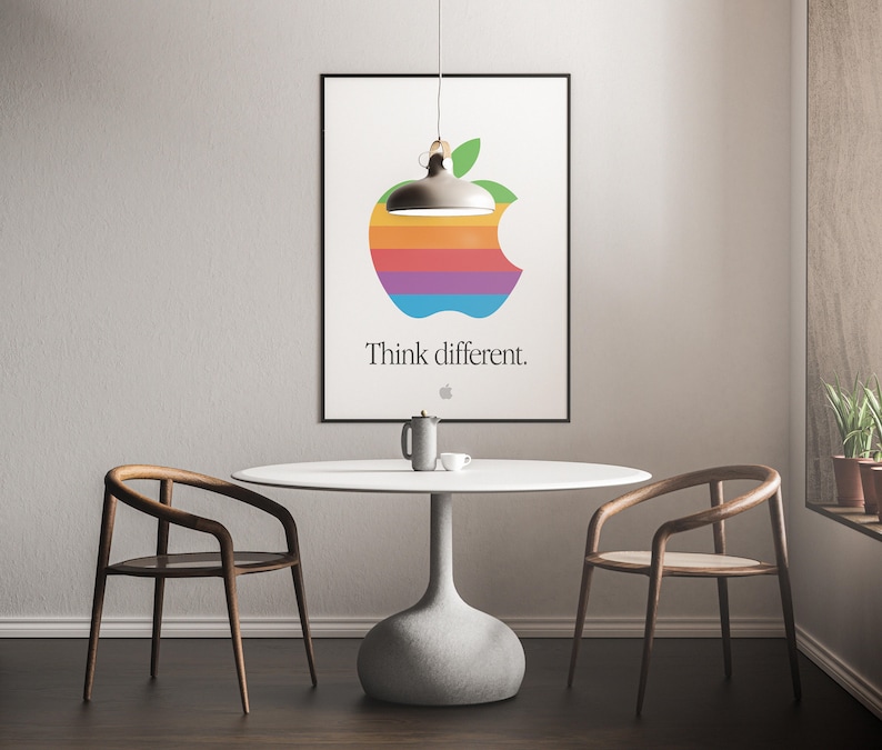 May include: A white poster with a rainbow apple logo and the text "Think different." hanging on a white wall above a white table with two wooden chairs.