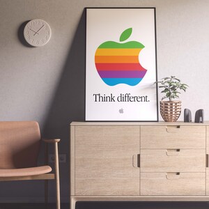May include: A framed poster with a rainbow apple logo and the text "Think different." The poster is hanging on a wall above a wooden cabinet with three drawers.