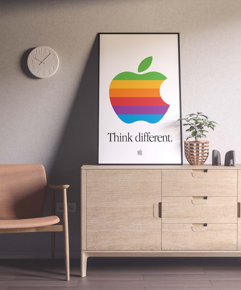 May include: A framed poster with a rainbow apple logo and the text "Think different." The poster is hanging on a wall above a wooden cabinet with three drawers.