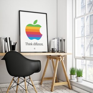 May include: A black framed poster with a rainbow colored Apple logo and the text "Think different." The poster is hanging on a white wall above a wooden desk with a black chair and a wooden easel.
