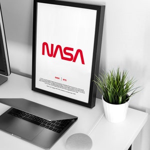 May include: A black framed poster with a red NASA logo and the text "NASA 1975" below it. The poster is on a white desk with a laptop, a potted plant, and a smart speaker.