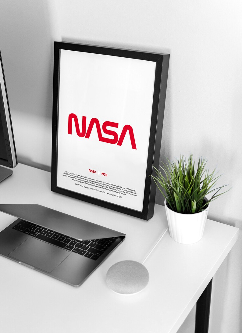 May include: A black framed poster with a red NASA logo and the text "NASA 1975" below it. The poster is on a white desk with a laptop, a potted plant, and a smart speaker.
