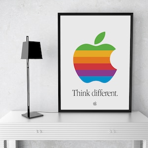 May include: A framed poster with a rainbow colored Apple logo and the text "Think different." The poster is hanging on a white wall above a white table with a black lamp.