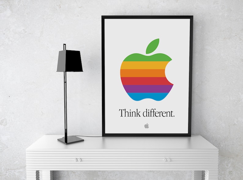 May include: A framed poster with a rainbow colored Apple logo and the text "Think different." The poster is hanging on a white wall above a white table with a black lamp.