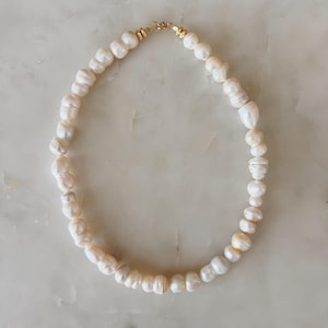 May include: A cream-colored pearl necklace with a gold clasp. The pearls are irregularly shaped and strung together to form a choker-style necklace. The necklace is displayed on a light-colored surface.