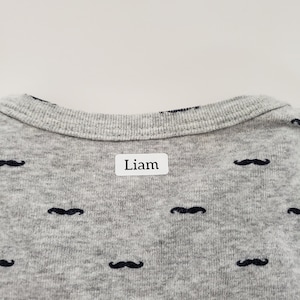 May include: Grey shirt with a black moustache pattern and a white label that says "Liam" on the back.