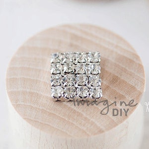 May include: A square-shaped rhinestone embellishment with a silver setting. The rhinestones are clear and arranged in a grid pattern.