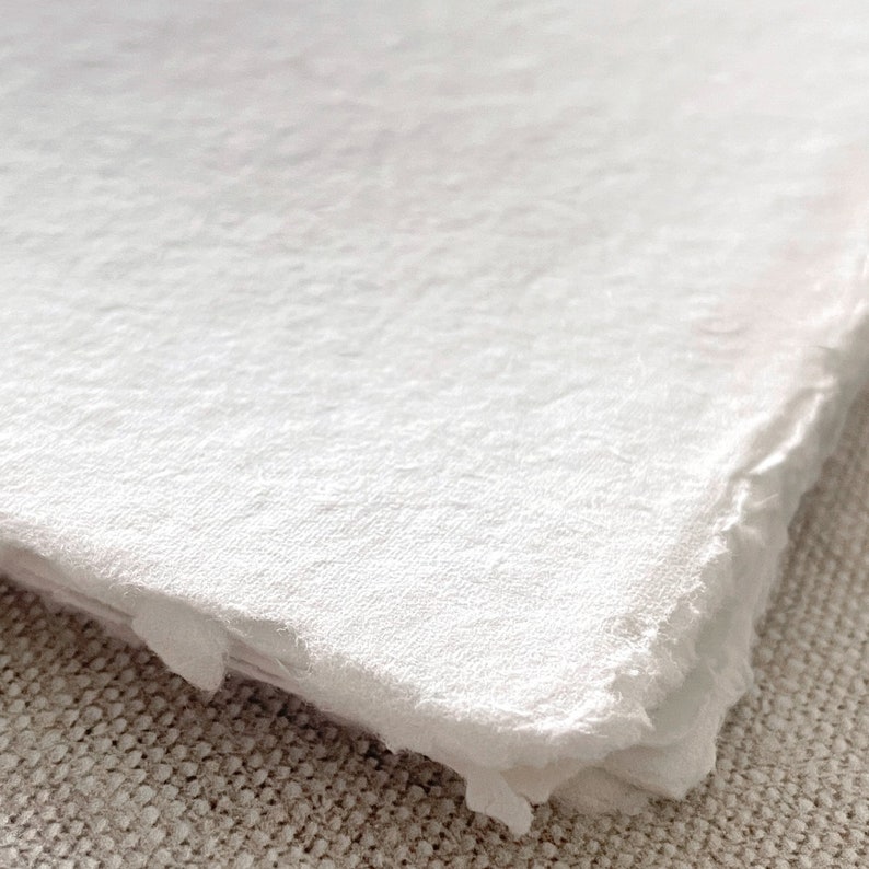 May include: A stack of white handmade paper with rough, deckled edges. The paper has a textured surface, giving it a natural, artisanal feel. The close-up view emphasizes the paper's texture and the soft, off-white hue.