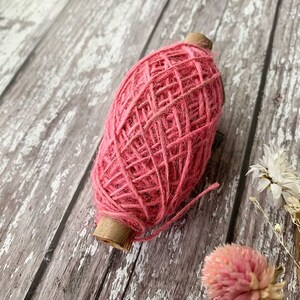 Könnte beinhalten: Eine Spule aus leuchtend rosa Garn, ideal für Bastelarbeiten und Dekorationen. Das Garn ist eng um einen Pappkern gewickelt und hat eine strukturierte Oberfläche. Der Hintergrund ist eine verwitterte Holzoberfläche.