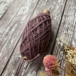 May include: A ball of mauve colored twine wound around a wooden spool. The twine is made of natural fibers and has a rustic look.