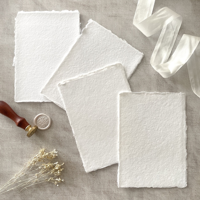 May include: Several handmade, textured white paper sheets with rough edges. A wooden handled wax stamp, a white wax seal, dried flowers, and a white ribbon are arranged around the paper. Suitable for stationery and invitations.