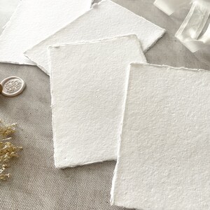 May include: Several handmade, textured white paper sheets with irregular edges. The paper is arranged in a layered composition, suggesting stationery or artistic use. A wax seal and ribbon are also visible.