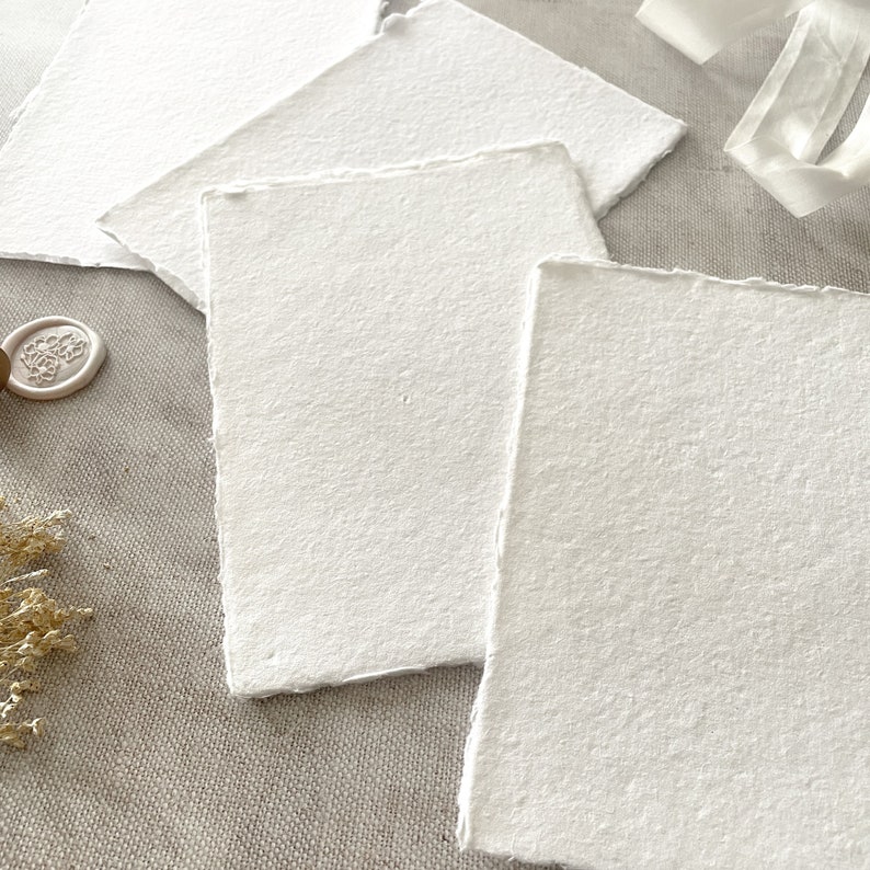 May include: Several handmade, textured white paper sheets with irregular edges. The paper is arranged in a layered composition, suggesting stationery or artistic use. A wax seal and ribbon are also visible.