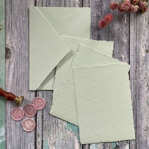 May include: A stack of five pale green paper sheets with deckled edges. The paper has a textured, handmade look. The sheets are arranged in a staggered pattern on a wooden surface.