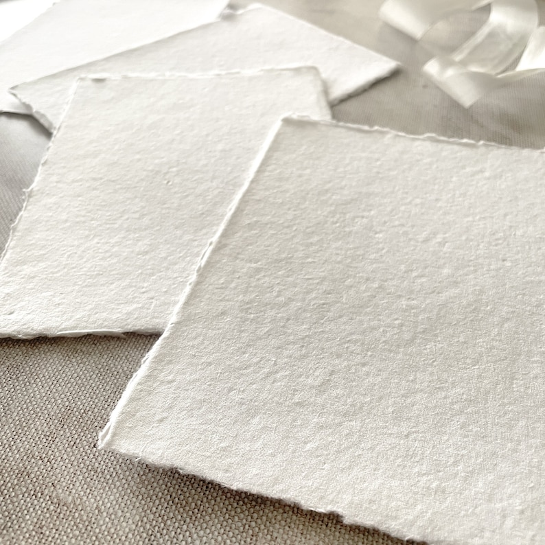 May include: Close-up of several handmade, textured white paper sheets with deckled edges. The paper is arranged on a neutral-toned surface, with a soft, natural aesthetic. The paper's surface has a slightly rough texture.