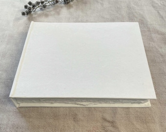 May include: A closed, rectangular, white book with a textured cover. The book is lying on a neutral-colored surface, with a sprig of dried flowers in the upper corner.