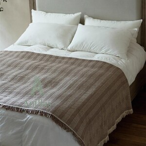 Pu&ograve; includere: Un letto con testiera grigio chiaro, cuscini bianchi e una coperta a righe marroni con frange. Il letto &egrave; fatto con lenzuola bianche e un piumino, creando una scena di camera da letto neutra e accogliente.