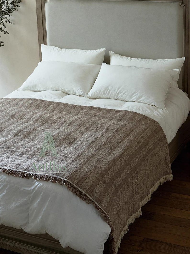 Pu&ograve; includere: Un letto con testiera grigio chiaro, cuscini bianchi e una coperta a righe marroni con frange. Il letto &egrave; fatto con lenzuola bianche e un piumino, creando una scena di camera da letto neutra e accogliente.