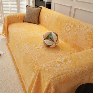 May include: A yellow sofa cover with a geometric pattern and fringe trim. A brown cushion rests on the sofa, and a decorative ball sits on the cover. The cover is draped over the sofa.
