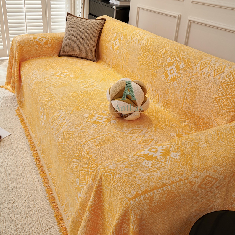 May include: A yellow sofa cover with a geometric pattern and fringe trim. A brown cushion rests on the sofa, and a decorative ball sits on the cover. The cover is draped over the sofa.
